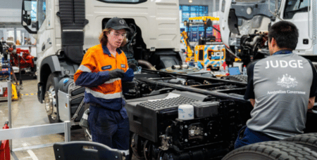 Photograph showing a technician in an orange and navy uniform explaining truck engine components to a judge wearing a gray shirt labeled "JUDGE Australian Government" inside a workshop. The setting includes large truck parts and tools, highlighting a technical evaluation or competition related to vehicle maintenance or repair.