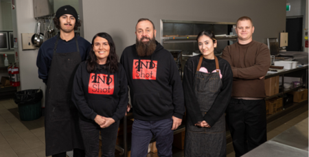Photograph of five people standing in a commercial kitchen, with three wearing aprons and two wearing black hoodies.