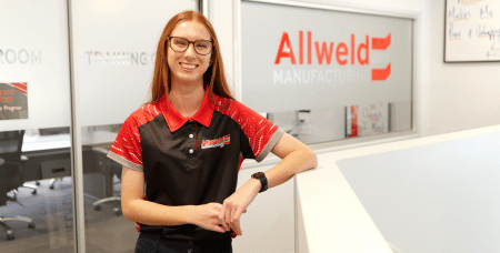 Photograph of a smiling woman wearing glasses and a black and red Allweld Manufacturing polo shirt standing inside an office with glass walls and company branding. The setting suggests a professional environment focused on manufacturing or training, with visible signage indicating rooms and company name.