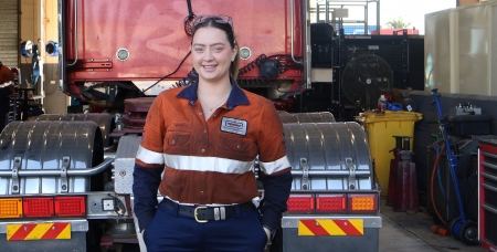 Image of Nicola Todd, smiling in orange high vis shirt and navy pants, standing in front of a truck in an automotive workshop