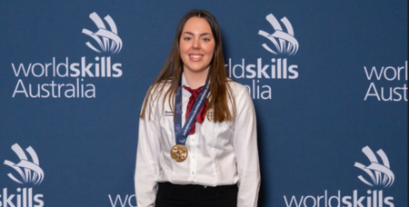Photograph of a young woman standing in front of a blue backdrop with "WorldSkills Australia" logos, wearing a white shirt, red tie, and a gold medal around her neck. The setting highlights her achievement in a skills competition, emphasizing recognition and success.