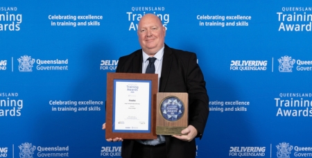 Image of a man in a suit and smiling at the camera, holding a trophy and certificate in front of a blue media wall