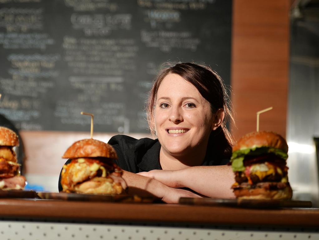 A woman stand at a counter with three burgers, behind her is a blackboard menu