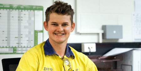 Photograph of a smiling man wearing a yellow high-visibility work shirt sitting in an office environment. Background includes a whiteboard with a calendar and office equipment, suggesting a professional or industrial workplace setting.