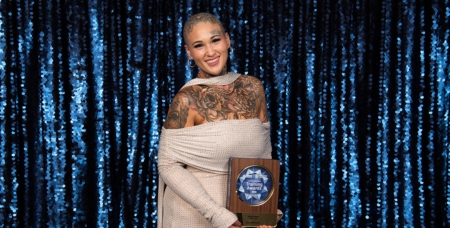 Image of Krystal Marie Lunny smiling in front of a blue bling wall holding her award trophy