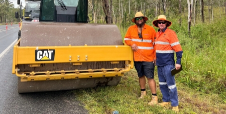 Image of two male workers on the side of a road in high vis gear near an asphalt roller machine