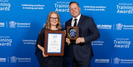 Image of a woman in a black dress holding a framed certificate and man in a suit holding a trophy, both smiling at the camera, in front of a blue media wall