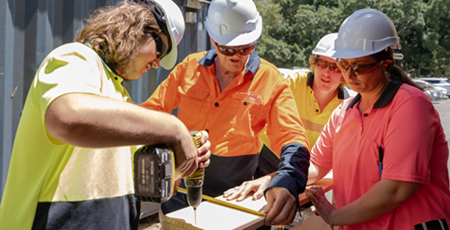 Man in hi-viz construction uniform holding a drill while three other watch on