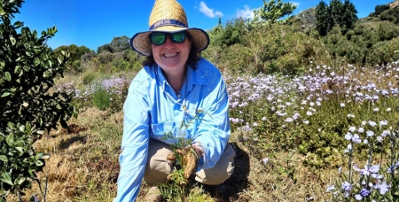 Image of Helaine Wilesmith in a blue shirt, hat and sunglasses, crouch im a garden and holding some plants in gloved hands while smiling at the camera