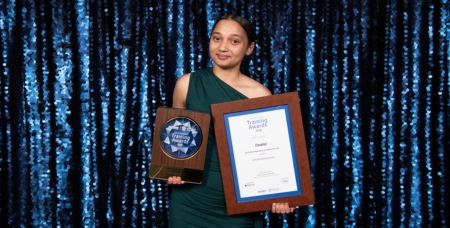 Image of Tahlia Mundraby-Holmes, young First Nations woman, smiling in front of a blue bling wall with her award trophy and certificate
