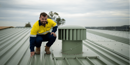 Photograph of a man wearing a yellow and navy work shirt crouching on a green metal rooftop next to a large cylindrical vent. The setting appears industrial or commercial with an overcast sky and trees in the background.
