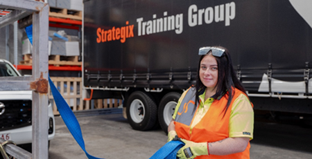 A woman wearing an orange safety vest and gloves holding a blue strap in a warehouse or loading area. A black truck with "Strategix Training Group" logo is visible in the background, indicating a training or instructional setting related to logistics or transportation.