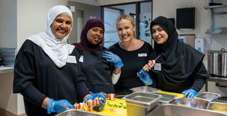 Four women in a commercial kitchen preparing food together, wearing gloves and aprons. They are smiling while assembling skewers with vegetables and meat at a food preparation bench