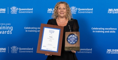 Image of Erin Pender, smiling in front of a blue media wall with her award trophy and certificate
