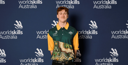 Photograph of a young man standing in front of a dark blue backdrop with repeated "WorldSkills Australia" logos in white. He is wearing a green and yellow collared shirt featuring an abstract design, representing Australia at a WorldSkills event.