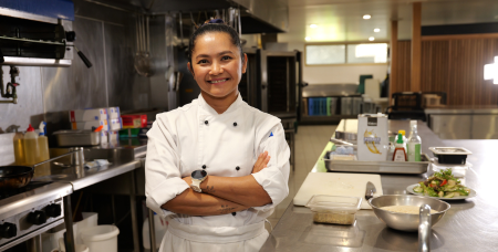 Photograph of a female chef standing confidently with arms crossed in a commercial kitchen. Kitchen features stainless steel counters, cooking equipment, and prepared dishes, highlighting a professional culinary environment.