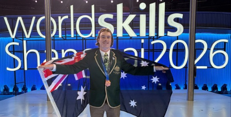 Image of Lachlan Wright in his Worldskills Australia uniform, drapped in the Australian flag and weariing his bronze medal in front of the WorldSkills Shanghai 2026 sign
